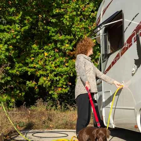 Frau füllt Wasser mit Wasserschlauch in ein Wohnmobil rein. Sie hat ein Hund an einer Leine