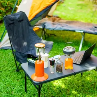Campingmöbel Ein Canpingplatz auf einer Wiese mit einm Stuhl einm Tisch mit einer Lampe und einem Kocher und einem Zelt im Hintergrund