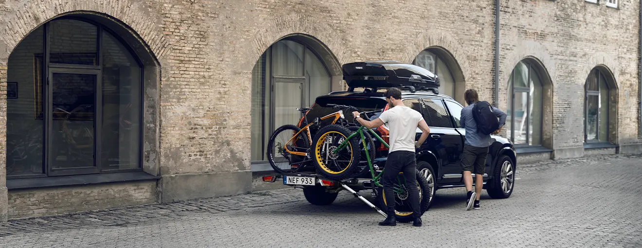 Zwei Personen beladen ihren Fahrradträger um eine Reise mit genügend Stauraum anzutreten, da eine Dachbox auf dem Auto montiert ist.